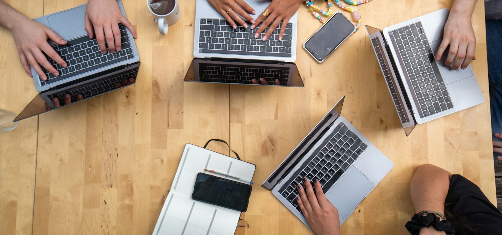 Foto mit 4 Notebooks auf einem Holztisch von oben. Jeweils Hände auf der Tastatur